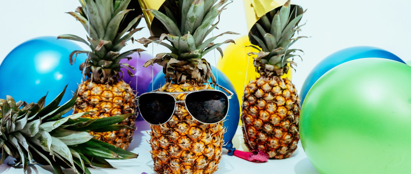 Ananas zwischen Luftballons mit Partyhüten und Sonnenbrille