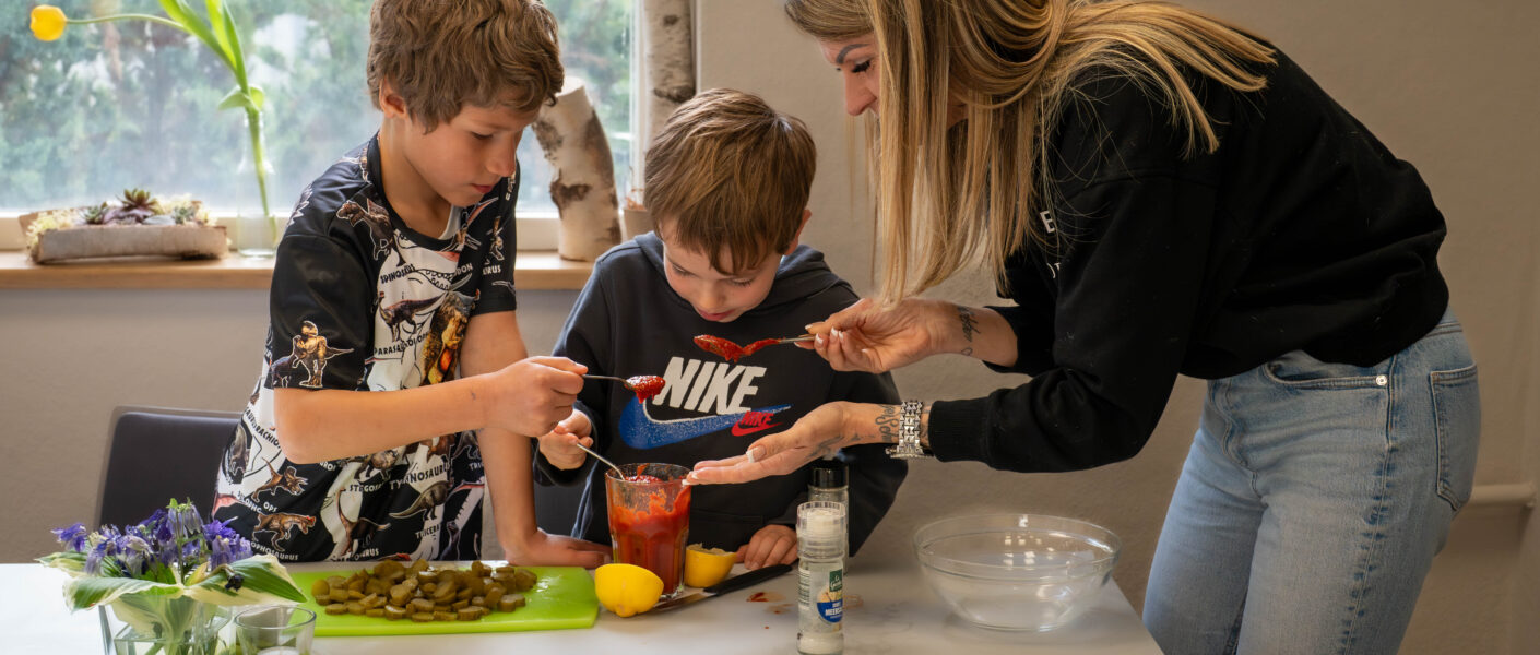 Frau kocht mit zwei Kindern am Tisch.